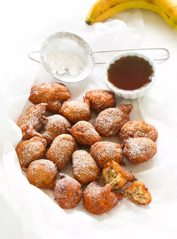 Image of Banana fritters dusted with icing sugar, with a strainer, banana and cup of maple syrup on a white background.