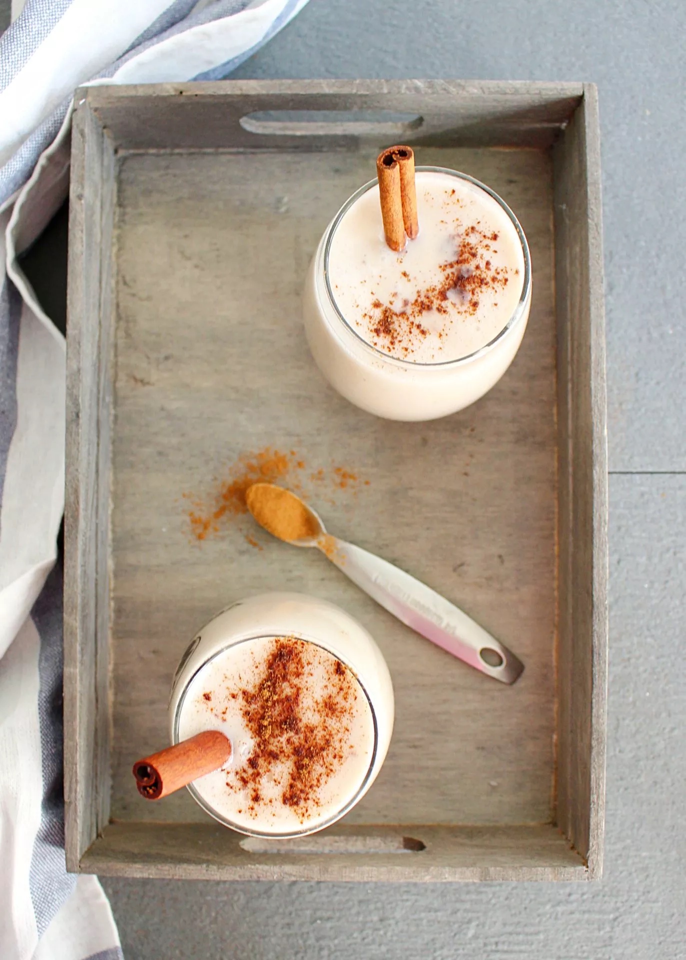 Image of two glasses on a wooden tray filled with a creamy white drink, decorated with cinnamon powder and cinnamon sticks.